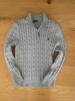 Grå stickad half zip-tröja Polo Ralph Lauren - Säljer en grå kabelstickad tröja från Polo Ralph Lauren med half zip och hög krage. Tröjan har lång ärm, ribbade muddar och den klassiska broderade loggan på bröstet. Perfekt för lager-på-lager under höst och vinter.