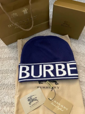 Blå mössa från Burberry - Säljer en snygg marinblå mössa från Burberry med vit logga runt hela kanten. Mössan är ribbstickad och har en klassisk passform. Perfekt accessoar för att lyfta din outfit och hålla dig varm under kyliga dagar. TILLKOMMER BARA MED DUSTBAG