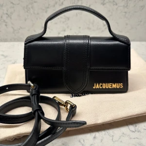 Svart Jacquemus handväska i läder - Fin svart läderväska från Jacquemus med guldfärgade detaljer. Modellen heter The Bambino och har måtten 17,5 x 9 cm. Axelbandet är reglerbart och avtagbart. Inköpt på Mytheresa 2022 för 450 EUR. Dustbag medföljer samt etiketter och kvitto 🖤