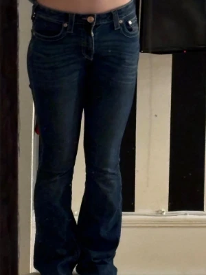 True Religion Becca Bootcut Jeans - Mörkblå jeans från True Religion, modell Becca Mid Rise Bootcut. Klassisk femficksdesign med snygga broderade detaljer på bakfickorna och bootcut-ben. Jeansen är i stretchigt denim och har en midwaist passform. Tajt vid låren och utsvängd längst ner