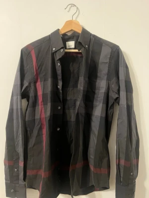 Svart rutig skjorta från Burberry - Snygg svart rutig skjorta från Burberry med klassiskt mönster i mörka toner och röda detaljer. Skjortan har lång ärm, knappar framtill och en stilren krage. Perfekt för dig som vill ha en ikonisk och trendig look.