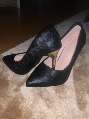 Svarta pumps med guldklack - Säljer ett par eleganta svarta pumps med spetsig tå och hög guldklack. Skorna har ett diskret blommigt mönster i materialet som ger en lyxig känsla. Insidan är ljusrosa och klacken är smal och metallisk. Perfekta för dig som vill sticka ut.