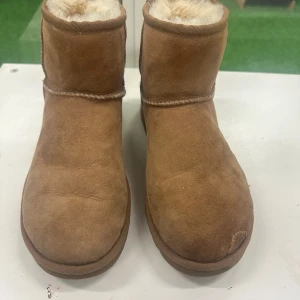 Bruna UGG boots i mocka 38 - Mysiga bruna UGG boots med rund tå och platt sula. Utsidan är i mjuk mocka och insidan är fodrad med fluffigt fårskinn för extra värme. Klassisk UGG-logga bak på hälen och enkel, stilren design. Perfekta för kalla dagar.( obs lite skada på vänster sida men inget förhinder)