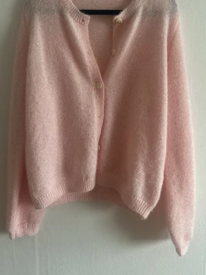 Ljusrosa stickad kofta med knappar - Supermjuk och fluffig ljusrosa stickad kofta med rund hals och vita knappar framtill. Koftan har långärmade ärmar och ribbade muddar vid hals, ärmslut och nederkant. Perfekt för lager-på-lager och chill dagar.