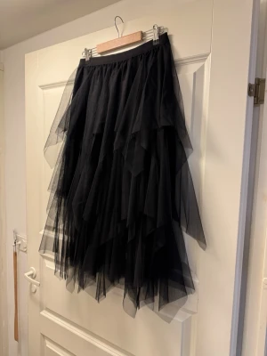 Svart långkjol i lager på lager-tulle M - Säljer en svart långkjol med flera lager av tunn, luftig tyll som ger en riktigt voluminös och cool look. Kjolen har elastisk midja och asymmetriska lager som skapar extra rörelse och attityd. Perfekt för dig som vill sticka ut med en edgy och trendig stil.