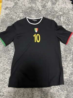Svart Dolce & Gabbana t-shirt med nummer 10 - Svart t-shirt från Dolce & Gabbana med guldfärgat nummer 10 på både fram- och baksida samt logga på bröstet. Kontrasterande vita, gröna och röda detaljer vid hals och ärmslut. Tillverkad i 100% bomull, perfekt för dig som gillar sportig stil och snygga detaljer.