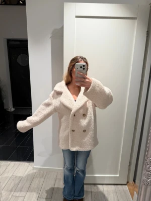 Beige teddyjacka med stora knappar - Supermysig teddyjacka i beige med stora slag och dubbelknäppning framtill. Jackan har en oversized passform och är tillverkad i fluffigt teddy-material som håller dig varm. Perfekt för dig som vill ha en trendig och bekväm look under kyliga dagar.