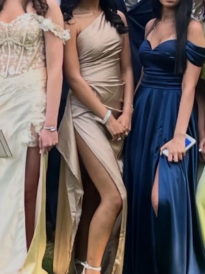 Champagnefärgad balklänning  - Säljer en champagnefärgad långklänning i glansigt satinliknande material. Klänningen har oneshoulder-design och en hög slits som ger en riktigt snygg siluett. Perfekt för dig som vill sticka ut på bal eller fest. Sitter figurnära och har en elegant drapering över höften.