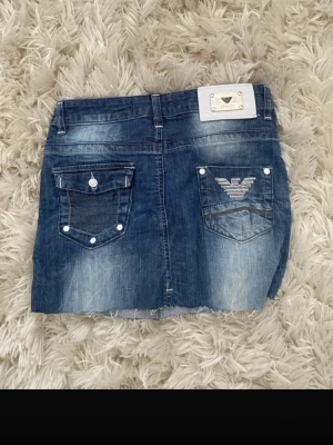 Emporio Armani jeanskjol med logga - Snygg blå jeanskjol från Emporio Armani med slitna detaljer, rå kant nertill och coola bakfickor med logga. Kjolen har vita detaljer vid fickorna och silverfärgade nitar. Perfekt för dig som vill ha en trendig och avslappnad look.