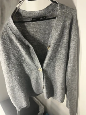 Grå stickad kofta från 157 - Mysig grå stickad kofta från 157 med v-ringning och två knappar framtill. Koftan har lång ärm och en enkel, klassisk design som passar perfekt till lager-på-lager. Skön och avslappnad passform.