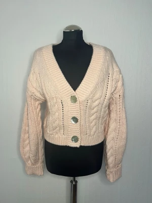 Ljusrosa stickad kofta från H&M - Supersöt ljusrosa stickad kofta från H&M i storlek XS. Koftan har grovstickat mönster med flätor, stora silvriga knappar framtill och bred ribbad kant. Croppad modell med långa ärmar och djup v-ringning. Perfekt för lager på lager under höst och vinter.