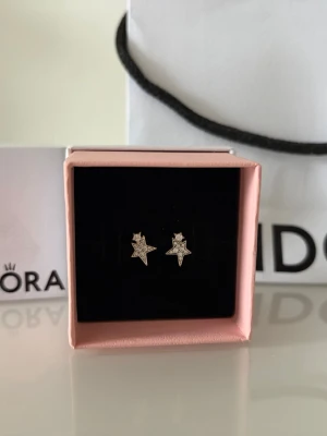 Pandora - Eleganta stjärnformade örhängen från Pandora, perfekta för att ge en gnistrande touch till vilken outfit som helst! De kommer i en snygg rosa smyckesask med svart sammetsfoder, tillsammans med den ursprungliga vita Pandora-asken och påsen – perfekt både som present eller för att unna dig själv. Helt nya, aldrig använda! Dessa örhängen är mångsidiga och passar både till vardags och till fest. Pris kan diskuteras, kom med förslag!