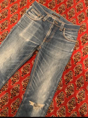 Blå Nudie Jeans med slitningar - Säljer ett par klassiska blå jeans från Nudie Jeans med snygga slitningar och en trasig detalj på ena benet. Jeansen har raka ben, fem fickOS och kontrastsömmar samt Nudie-logga på bakfickan. Perfekt för dig som gillar en avslappnad och trendig look.
