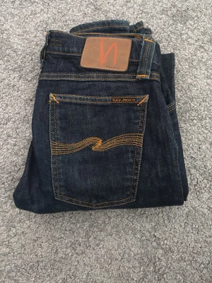 Mörkblå jeans från Nudie Jeans Co - Snygga mörkblå jeans från Nudie Jeans Co med klassiska orange sömmar och broderad bakficka. Jeansen har en rak passform och är tillverkade i slitstarkt denim. Perfekta för dig som gillar stilrena och tidlösa jeans.