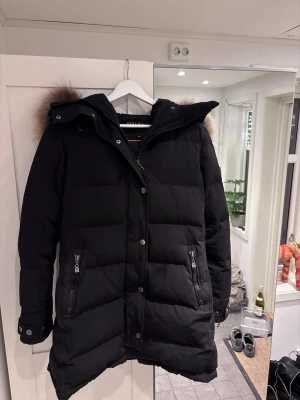 Svart lång pufferjacka Hollies - Säljer en svart lång pufferjacka från Hollies med stor luva och fluffig pälsdetalj i brunt runt huvan. Jackan har dragkedja och knappar framtill, två fickor med dragkedja och snygga knappdetaljer på utkanterna. Perfekt för kalla dagar! Pris kan diskuteras 