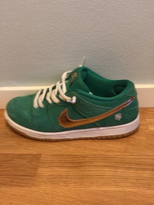 Nike Dunk Low SB St. Patrick's Day - Gröna Nike Dunk Low SB med guldiga swoosh-detaljer och vit sula. Skorna har en fyrklöver broderad på sidan och vita skosnören. Ovandelen är i mocka och insidan är fodrad i grönt tyg. Perfekt för dig som vill sticka ut med en unik sneaker. Ingen skolåda! Ny pris nya med sko låda i denna storlek 3500 kr andvända helt ok mycket storlek 42.