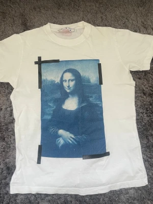 Vit Off-White t-shirt med Mona Lisa-tryck - Vit t-shirt från Off-White med stort blått Mona Lisa-tryck framtill och svarta tejpdetaljer runt motivet. Klassisk passform med rund hals och korta ärmar. Diskret Off-White-logga i nacken. Perfekt statement-plagg för dig som gillar streetwear.