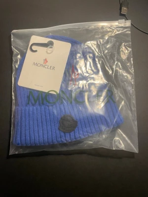 Blå Moncler mössa i ribbstickad ull - Snygg blå mössa från Moncler i ribbstickad ull. Mössan har en uppvikt kant och en svart Moncler-logga framtill. Perfekt accessoar