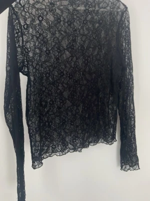 Svart spetsblus från Gina Tricot - Säljer en svart långärmad spetsblus från Gina Tricot. Blusen är helt genombruten med blommigt spetsmönster och har en tight passform. Perfekt att styla med ett linne under eller som layering-piece. Storlek L men passar mer allt från xs-M