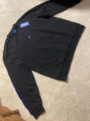 Svart sweatshirt från Hugo med blå detaljer - Säljer en oanvänd och ny Hugo boss tröja med etiketten kvar. Är i storlek M men är stor i passformen 