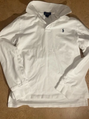 Vit långärmad henley från Polo Ralph Lauren - Klassisk vit långärmad henley från Polo Ralph Lauren med blå broderad logga på bröstet. Skjortan har en mjuk krage och är tillverkad i bomull för en skön känsla. Perfekt till jeans eller chinos för en clean och stilren look.