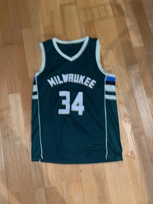 Milwaukee Bucks Antetokounmpo tröja - Säljer en mörkgrön Milwaukee Bucks baskettröja med nummer 34 och namnet Antetokounmpo på ryggen. Tröjan har vita och blå detaljer på ärmarna och vita kanter runt hals och ärmhål. Perfekt för basketfans som vill sticka ut.