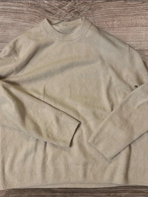 stickad tröja Arket - Säljer en stilren beige stickad tröja i storlek M. Tröjan har rund halsringning, ribbade muddar och är långärmad. Perfekt för lager-på-lager och passar till många olika outfits.