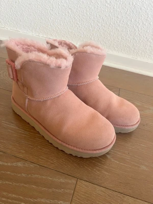 Rosa UGG - Mysiga rosa UGG boots med fluffigt foder och dekorativt spänne på sidan. Tillverkade i mjuk mocka med beige sula och rund tå. Perfekta för kalla dagar när du vill ha både stil och värme.