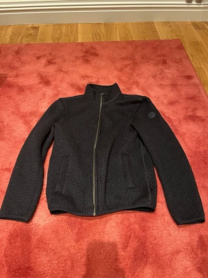 Svart stickad jacka från Colmar - Säljer en svart stickad jacka från Colmar med dragkedja framtill och hög krage. Jackan har två fickor och en rund Colmar-logga på ärmen. Perfekt för kyliga dagar och har en clean, stilren look som funkar till det mesta.