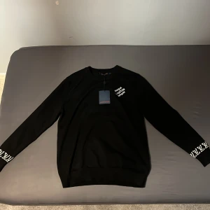 Svart Louis Vuitton sweatshirt - Svart sweatshirt från Louis Vuitton med vit broderad text 'Marque L Vuitton Deposee' på bröstet och logotypdetaljer på ärmsluten. Tröjan har rund hals och långa ärmar, tillverkad i mjuk bomull för en skön känsla. Perfekt för dig som gillar exklusiva streetwear-vibbar.