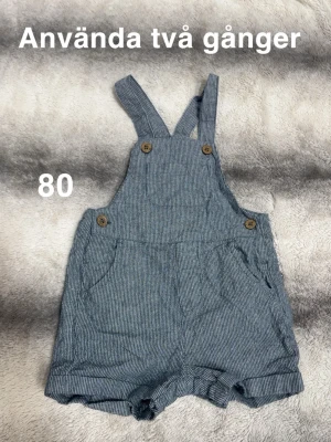  blå byxdress med knappar - Supersöt byxdress med hängslen och träknappar framtill. Byxdressen har korta ben och praktiska fickor på sidorna. Perfekt för varma dagar och enkel att matcha med en t-shirt under. 🌸 Använda två gång. Storlek 80