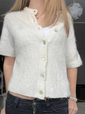 Vit stickad kofta från Gina Tricot - Superfin vit stickad kofta från Gina Tricot med korta ärmar och stora knappar framtill. Koftan är ribbstickad och har en lite boxig, croppad passform som är perfekt till jeans eller kjol. Enkel och stilren design som funkar året runt.