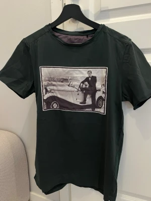 Mörkgrön t-shirt Limitato med fototryck - Cool mörkgrön t-shirt från Limitato med ett stort fototryck på bröstet som visar en man vid en klassisk bil. T-shirten har rund hals och är tillverkad i mjuk bomull. Perfekt för dig som gillar unika prints och streetstyle. Sömmen har släppt halva baktill längst ner samt på ena armen längst ut (se bilder)