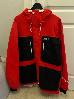 Röd och svart skidjacka från CLWR - Säljer en röd och svart skidjacka från CLWR med stora fickor framtill, vita dragkedjor och huva med snörning. Jackan har beige quiltat foder och coola detaljer som logga på bröstet och ryggen. Perfekt för dig som vill sticka ut i höst, vinter och ute i backen!