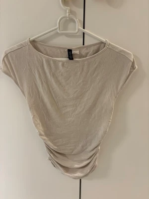 Beige topp från H&M Divided - En enkel  topp från H&M Divided. Modellen är ärmlös med breda axlar och har en lätt draperad effekt i sidorna. Tunt och mjukt material som känns skönt mot huden. 