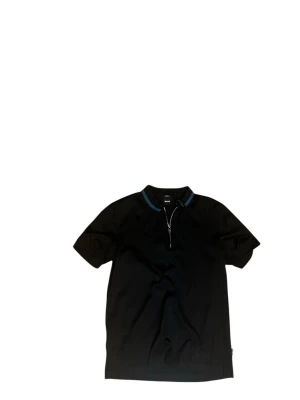 Svart Piké från BOSS - Svart piké från BOSS med slim fit passform och korta ärmar. Pikén har en snygg krage med blå detalj och en halv dragkedja framtill. Tillverkad i merceriserad bomull som ger en mjuk och lyxig känsla. Perfekt för dig som gillar stilrena och moderna plagg. Passar S/M