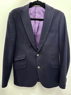 Mörkblå kavaj från Morris, stl 46 - Snygg mörkblå kavaj från Morris med lila foder och dekorativa knappar. Klassisk dubbelknäppt modell med tre fickor framtill. Tillverkad i en mix av ull, polyamid och kashmir för en lyxig känsla. Försiktigt använd, som ny!