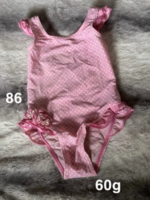 Rosa prickig baddräkt med volanger - Söt rosa baddräkt med vita prickar och volangdetaljer vid axlar och ben. Dekorerad med små blommor på ena höften. Perfekt för strand eller poolhäng. 🌸 Fint använt skick. Storlek 86