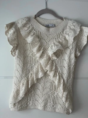 Beige spetsstickad topp från Zara - Supersöt beige topp från Zara i stickad bomull med spetsvolanger över axlar och framtill. Kortärmad modell med rund halsringning och detaljerad hålstickning. Perfekt för dig som gillar romantiska och feminina detaljer.