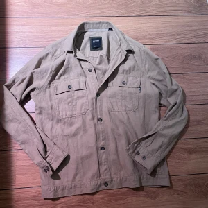 Beige overshirt från Only & Sons - Snygg beige overshirt från Only & Sons med klassisk krage, två bröstfickor med knappar och långa ärmar. Perfekt lager-på-lager-plagg med avslappnad passform och stilrena detaljer. Stängs med knappar framtill.