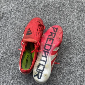 Adidas Predator röda fotbollsskor - Säljer ett par Adidas Predator fotbollsskor i rött med vita och svarta detaljer. Skorna har snörning, mönstrad ovandel och tydlig Predator-logga på sulan. Perfekta för dig som vill sticka ut på planen med snygg design och bra grepp.