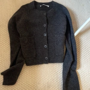 Svart kofta med knappar - Mysig svart kofta med rund hals och stora knappar framtill. Tröjan har långa ärmar och två praktiska fickor på framsidan. Perfekt för att hålla sig varm och samtidigt se chill ut. Passar dig som gillar enkel och stilren design.