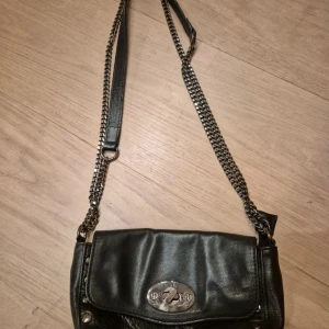 Svart axelväska - Snygg svart axelväska i skinn från Hallhuber med kedjerem och coolt vridlås i silver. Väskan har ett huvudfack med dragkedja och innerficka, perfekt för mobilen och småprylar. Stilren design som lyfter vilken outfit som helst.