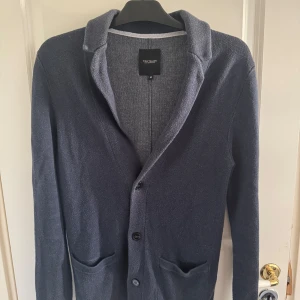 Mörkblå kofta från Tom Tailor - Snygg mörkblå kofta från Tom Tailor med klassisk krage och knäppning framtill. Koftan har två praktiska fickor och är tillverkad i ett mjukt stickat material som passar perfekt till lager-på-lager-stil. Enkel att matcha med både jeans och chinos.