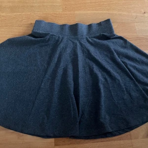 Mörkgrå kortkjol från H&M Divided - Mörkgrå kortkjol från H&M Divided med bred resår i midjan. Kjolen är tillverkad i en mix av polyester, bomull och elastan.