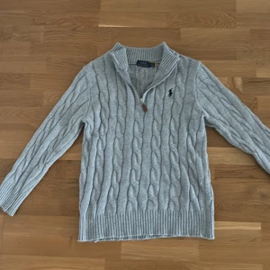 Grå stickad half zip tröja Polo Ralph Lauren - Snygg grå kabelstickad tröja från Polo Ralph Lauren med half zip och ribbad krage. Tröjan har långärmade ärmar och klassisk svart Polo-logga broderad på bröstet. Perfekt för dig som gillar stilren och tidlös design.