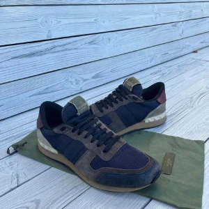 Valentino Rockrunner  - Snygga Valentino Rockrunner sneakers i mörkblått tyg med grå och beige mockadetaljer samt en vinröd häl. Skorna har klassisk snörning och en platt sula med gummidetaljer. Perfekta för dig som vill ha en stilren och exklusiv look.