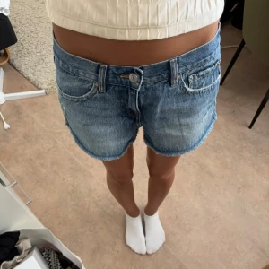 Blå jeansshorts från Gina Tricot - Snygga blå jeansshorts från Gina Tricot med klassisk femficksdesign och rå kant nedtill. Modellen är midwaist och har en avslappnad passform. Perfekta för sommaren och lätta att matcha med en crop top eller hoodie.