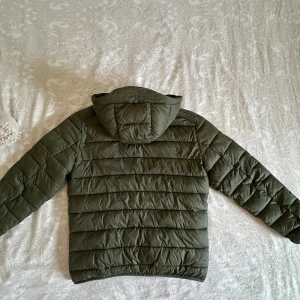 Olivgrön pufferjacka från Lindbergh - Säljer en olivgrön pufferjacka från Lindbergh med huva och dragkedja. Jackan är quiltad och har en clean look med diskret logga på ärmen. Perfekt för kyliga dagar och enkel att matcha med olika outfits.