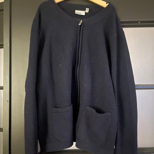 Mörkblå kofta från Isolde med dragkedja - Säljer en mörkblå kofta från Isolde i storlek M. Koftan har ett diskret strukturmönster, två praktiska fickor framtill och stängs med dragkedja. Perfekt för lager-på-lager och passar till många olika outfits.